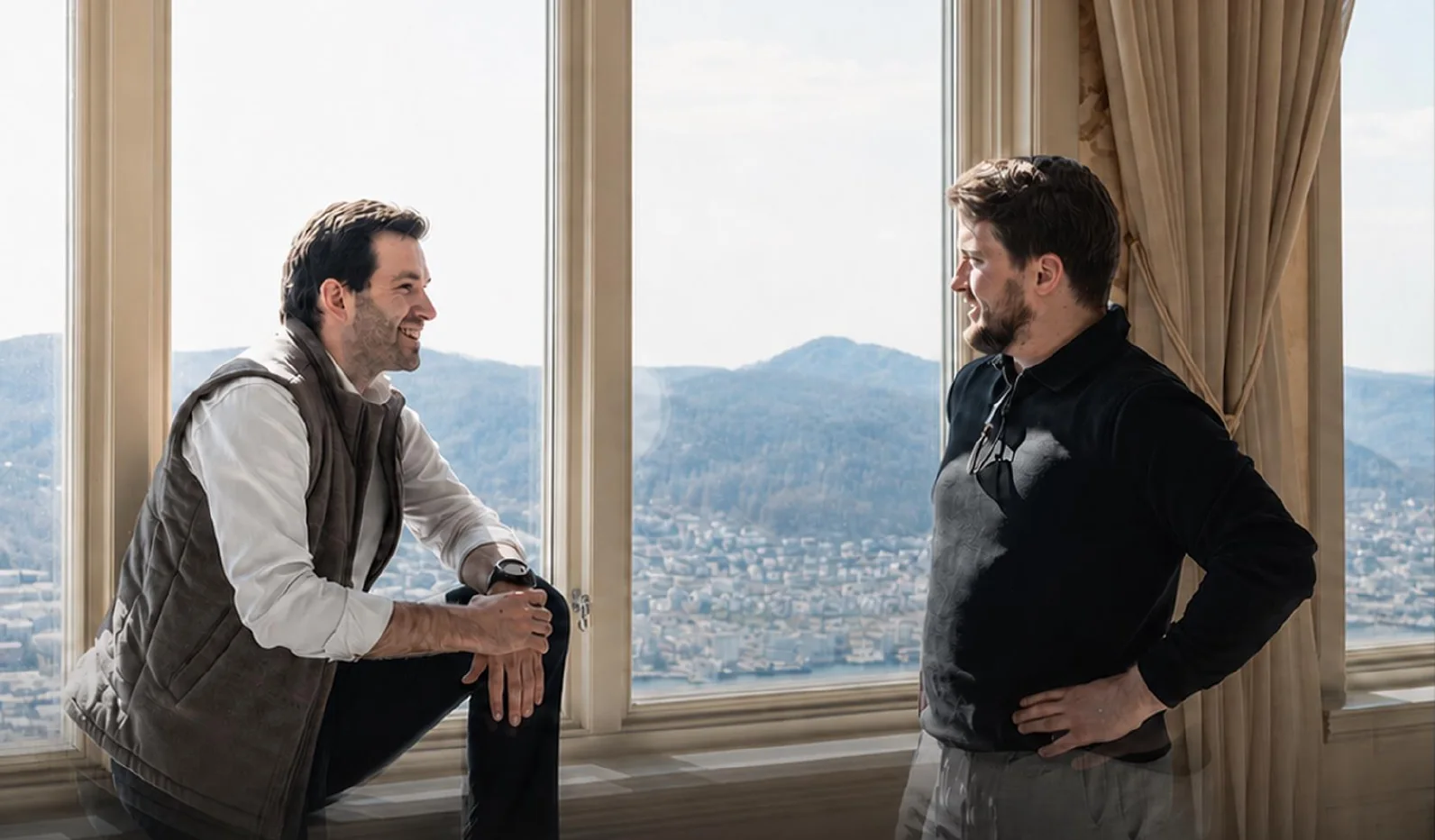 Njål Eliasson sitting on a window ledge and Mathias Haugsbø standing beside him, talking, with Bergen behind the window